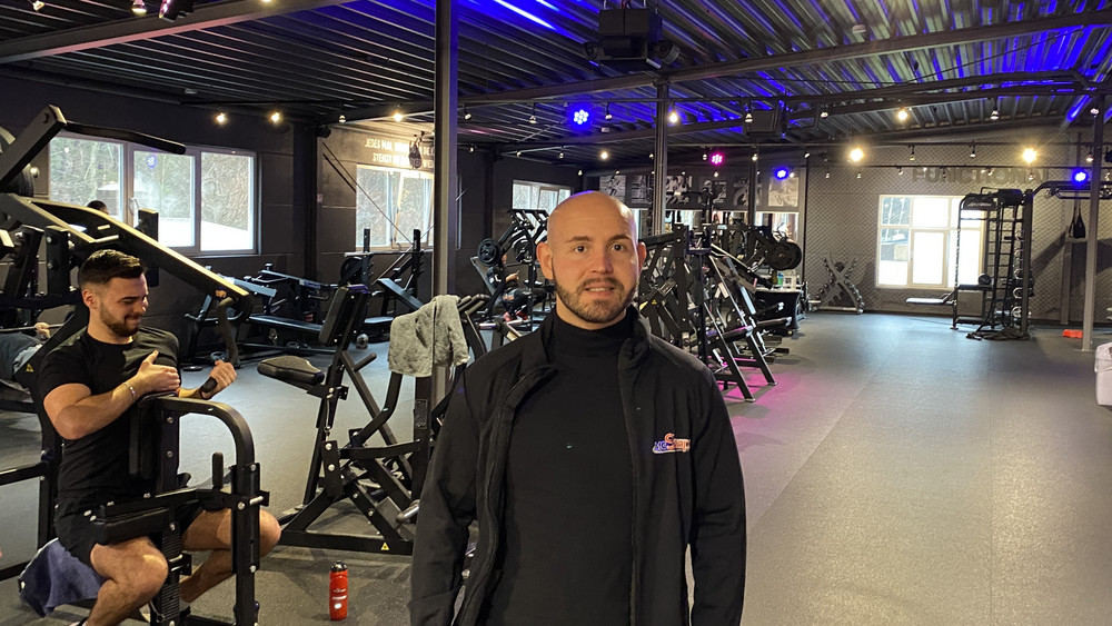 Sascha Mössinger steht in seinem Fitnessstudio in Stadtallendorf. Im Hintergrund trainiert an Mann einem Kraftgerät.