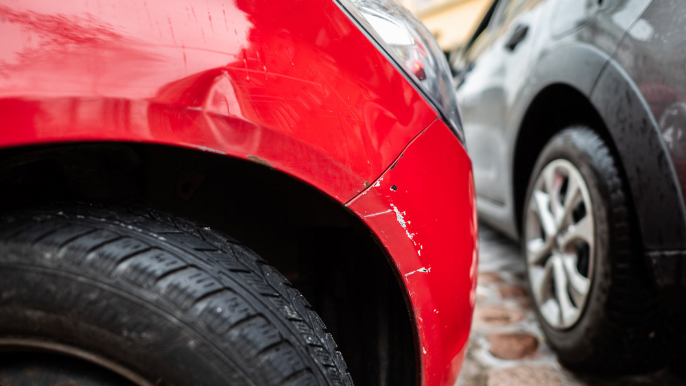 Ein rotes Autos steht mit einem Blechschaden in einer Parklücke, während ein graues Auto vorbei fährt.