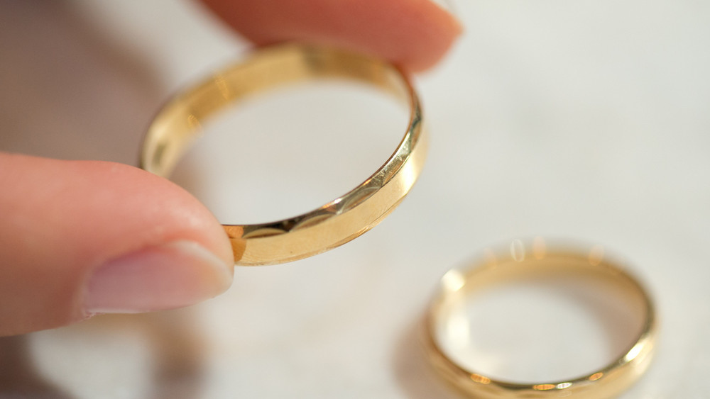 Eine Person hält einen goldenen Ehering in der Hand. Daneben liegt ein weiterer Ring.
