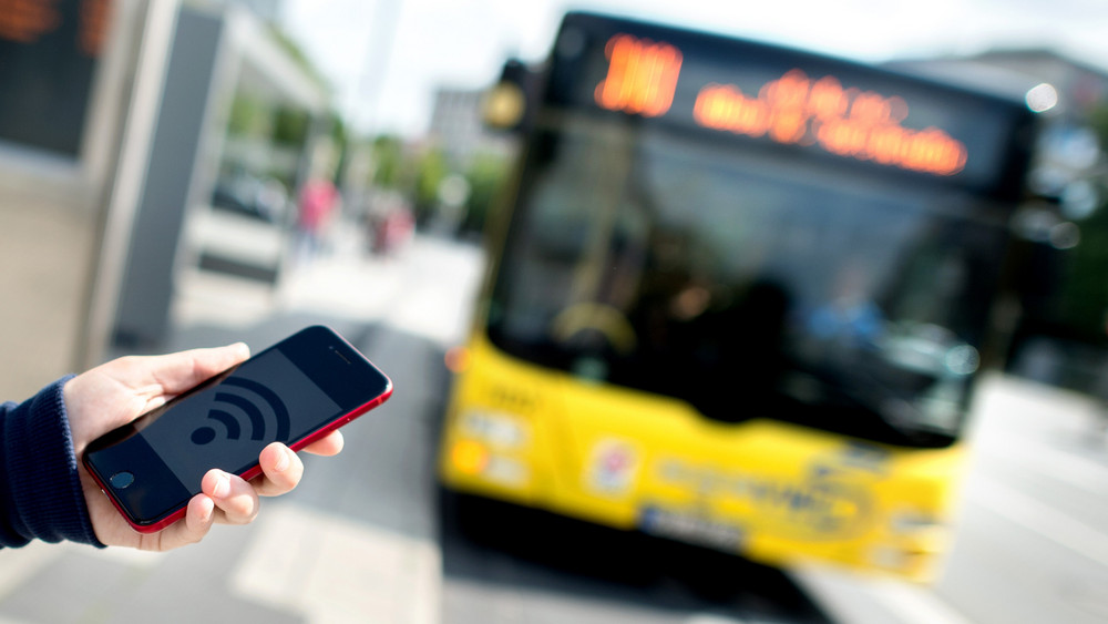 Jemand hält ein Handy mit einem WLAN-Symbol und im Hintergrund sieht man einen Bus