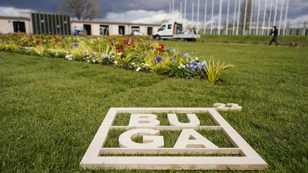 Ein Holzrahmen mit der Aufschrift „BUGA 23“ liegt auf dem Gelände der Bundesgartenschau vor einem Blumenbeet.