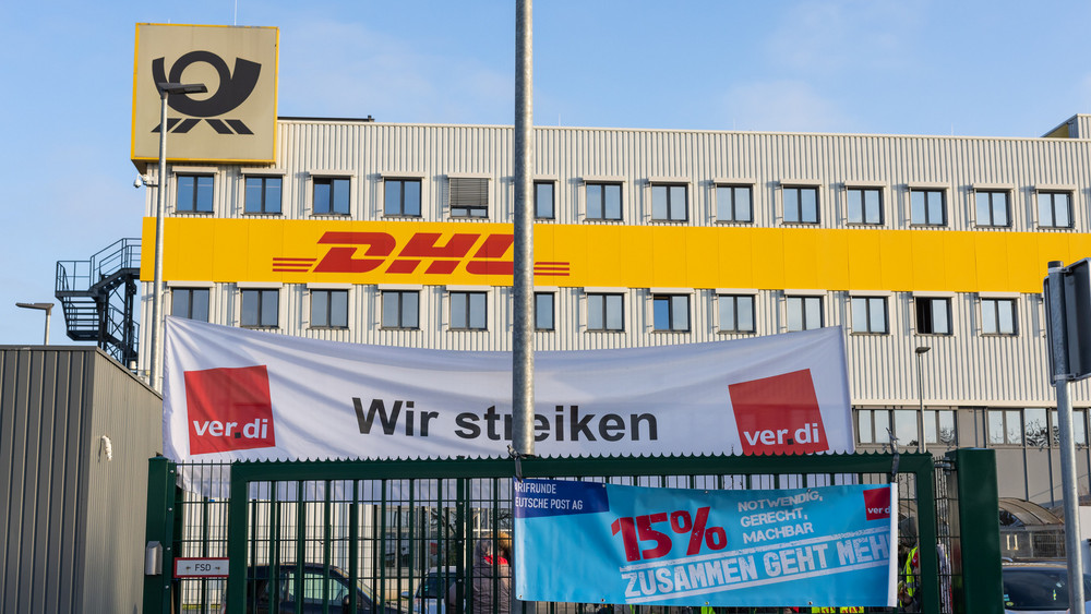 Ein Banner mit der Aufschrift "Wir streiken" der Gewerkschaft Ver.di hängt vor einem Gebäude des Paketdienstleisters DHL.