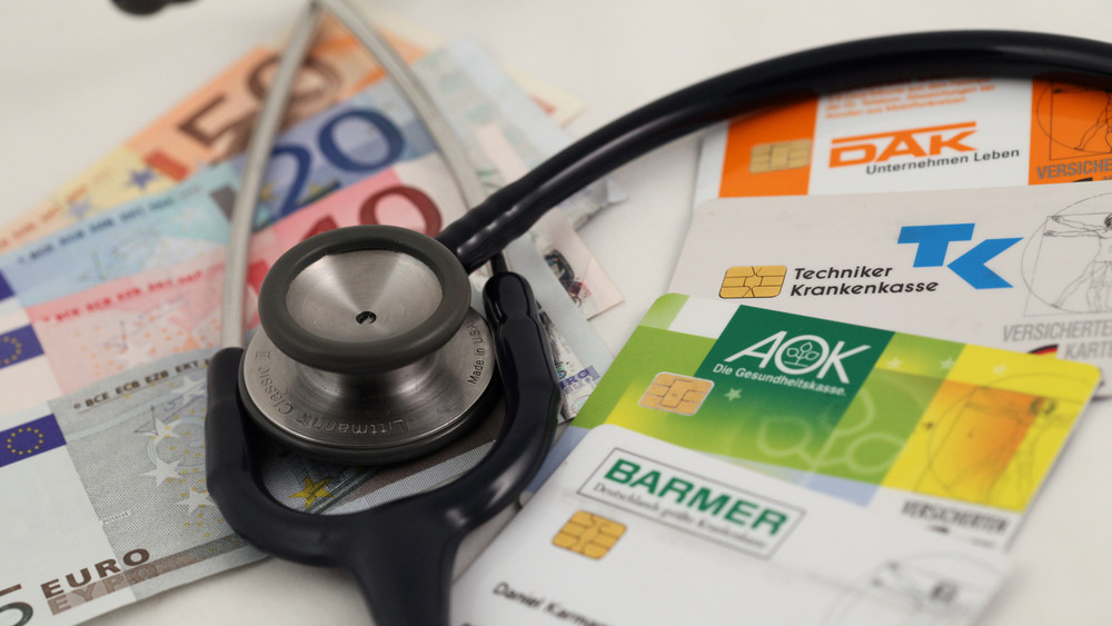 Die Versichertenkarten der deutschen Krankenkassen DAK, AOK, Barmer und Techniker Krankenkasse TK liegen mit einem 50, 20, zehn und fünf Euro-Schein unter einem Stethoskop.