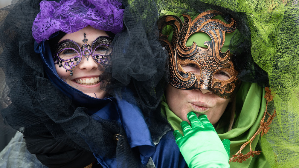 Zwei Närrinnen mit venezianischen Masken lachen in die Kamera. In Lila und Grün stürzen sie sich in den Faschings-Trubel.