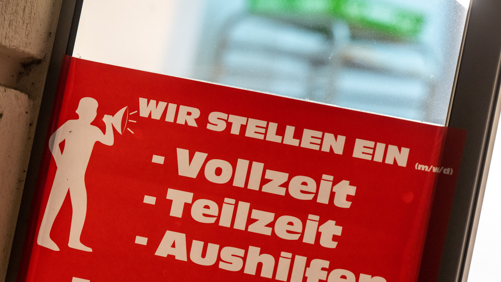 Ein rotes Schild mit der Aufschrift „Wir stellen ein - Vollzeit, Teilzeit, Aushilfen„ hängt am Eingang einer Bäckerei (Archivbild).