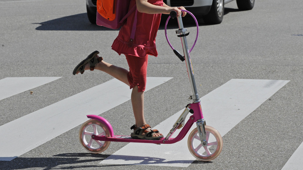 Ein junges Mädchen fährt mit einem Tretroller über einen Zebrastreifen.