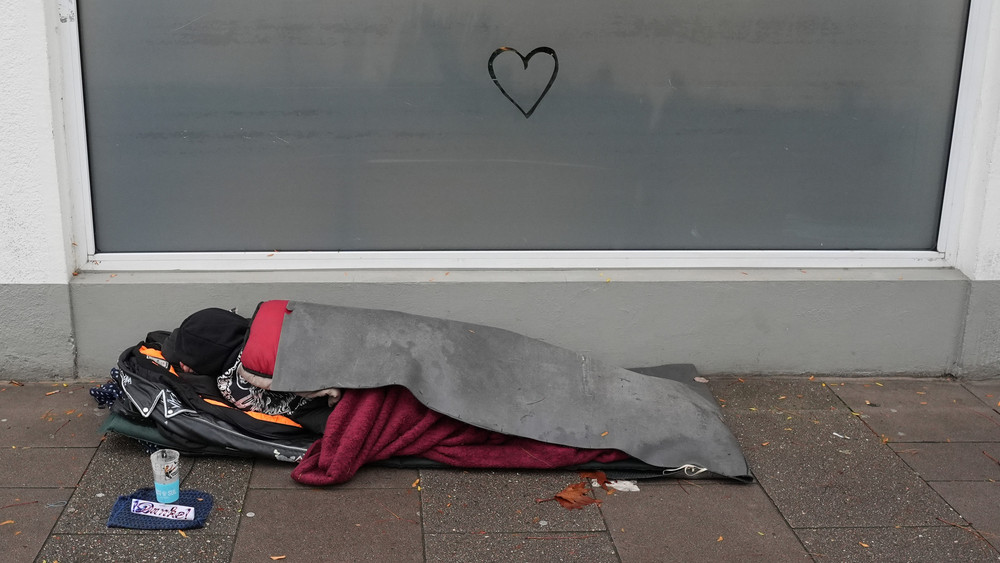 Ein obdachloser Mensch liegt in einem Schlafsack auf dem Gehweg vor einem Supermarkt.