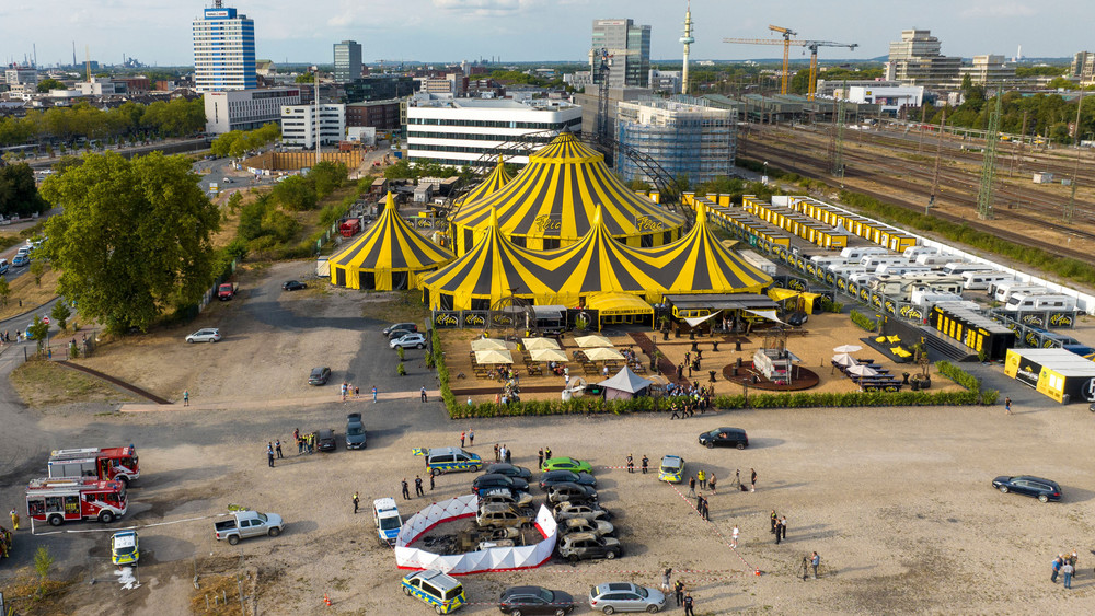 Ausgebrannte Fahrzeuge stehen nach dem Absturz eines Kleinflugzeugs an dem aufgestellten Sichtschutz, mit dem das Flugzeugwrack abgeschirmt wurde. Im Hintergrund ist das Zelt des Zirkus FlicFlac in Duisburg zu sehen. 