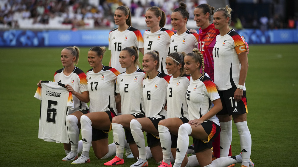 Vor dem Olympia-Auftaktspiel gegen Australien nehmen die deutschen Spielerinnen Aufstellung für ein Mannschaftsfoto. Dabei halten sie ein Trikot der verletzten Mitspielerin Lena Oberdorf in die Höhe.