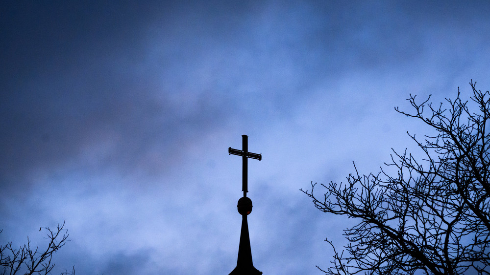 Dunkle Wolken ziehen über das Kreuz auf einer evangelischen Kirche hinweg.