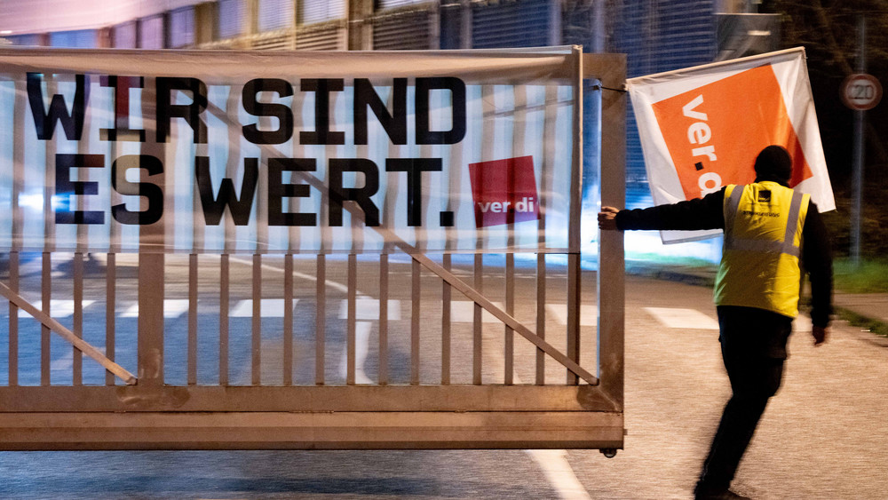 Ein Verdi-Mitglied schließt ein Werkstor an dem ein Poster mit der Aufschrift "Wir sind es wert." hängt.