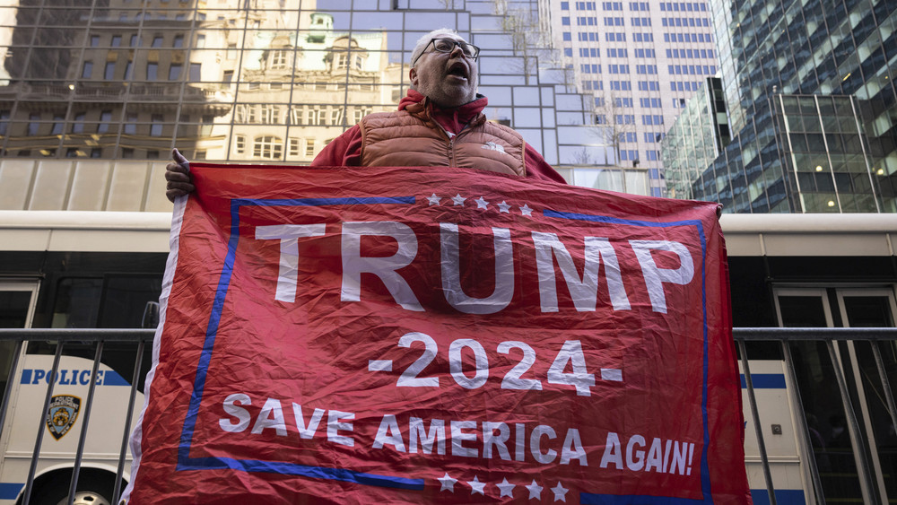 Ein Unterstützer des ehemaligen Präsidenten Trump hält vor dem Trump Tower eine Fahne mit der Aufschrift «Trump 2024 - Rette Amerika noch einmal». Ex-US-Präsident Trump wird nach seiner Anklage vor Gericht erwartet. Er selbst inszeniert sich als Opfer einer politisch gesteuerten Justiz. Erste Proteste sind bereits angekündigt.