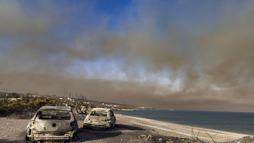 In der Ortschaft Kiotari stehen zwei ausgebrannte PKWs am Strand, am Himmel treibt der Wind eine riesige Rauchwolke in Richtung Nordosten.
