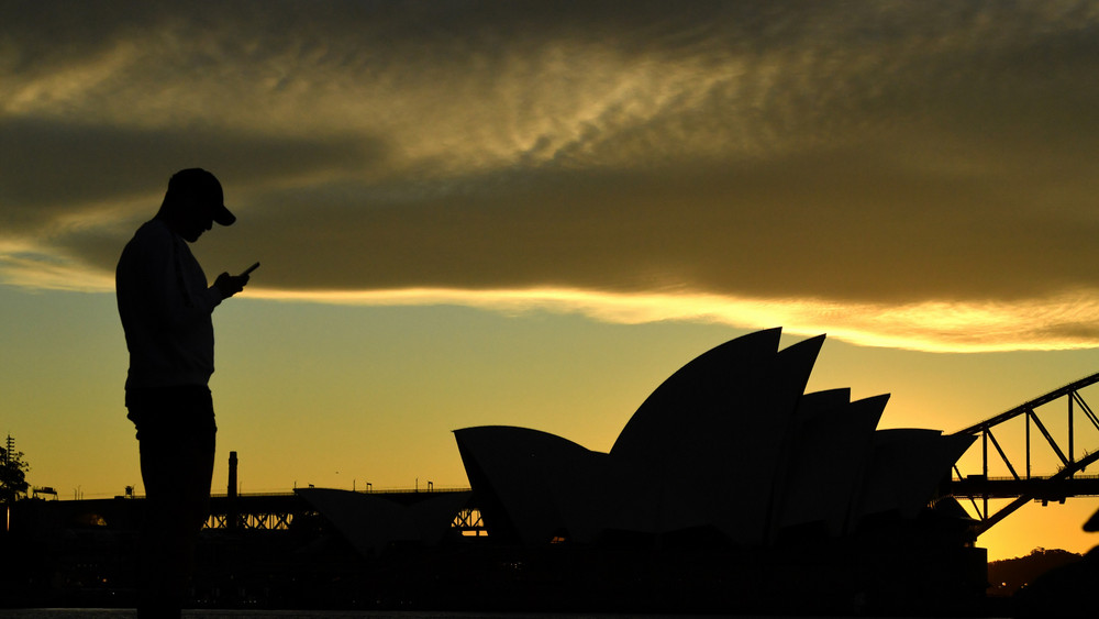 Ein Mann steht bei Sonnenuntergang vor dem Sydney Opera House und blickt auf sein Mobiltelefon. In Australien hat ein Totalausfall beim zweitgrößten Mobilfunkanbieter Optus die Telefon- und Internetverbindungen von mehr als zehn Millionen Menschen lahmgelegt (Symbolbild).