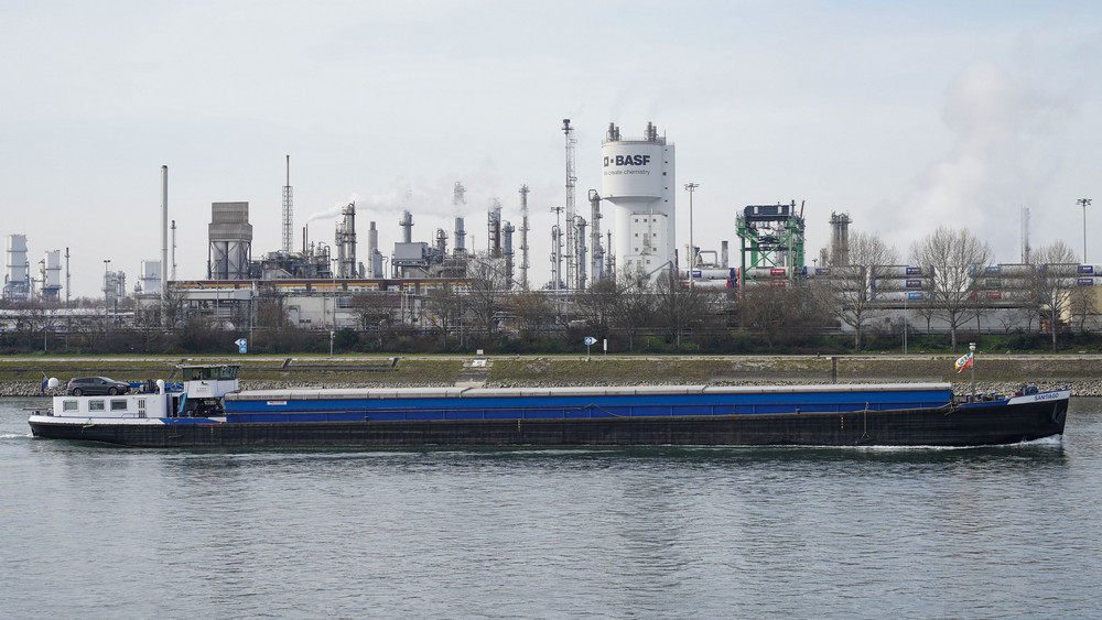 Ein Turm mit der Aufschrift „BASF“ steht neben Schornsteinen auf dem Werksgelände des Chemiekonzerns BASF. Im Vordergrund fährt ein Schiff über den Rhein