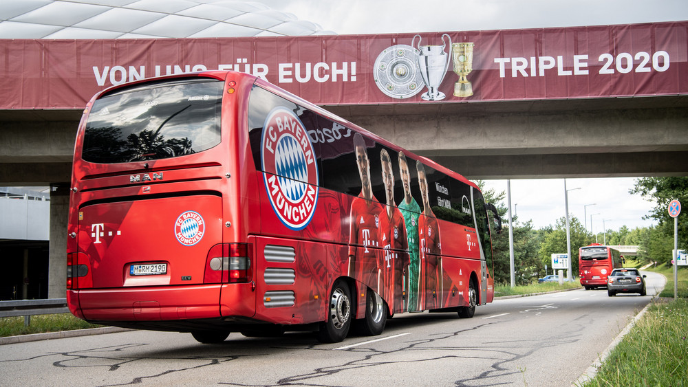 Die Spieler vom FC Bayern München fahren mit ihren Mannschaftsbussen unter einem Banner mit der Aufschrift "Von uns für euch! Triple 2020" vor der Allianz Arena hindurch.