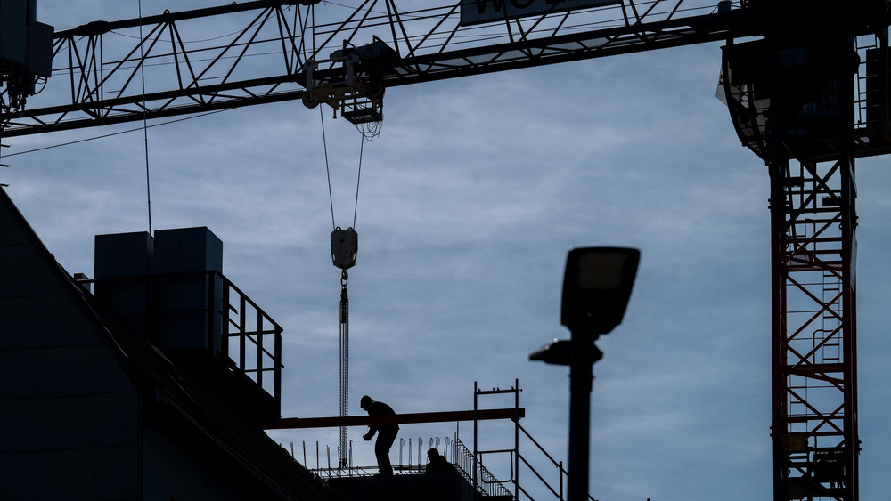 Ein Symbolbild zur Baubranche. Es zeigt eine Person auf einem Gerüst, eine Baustelle und Kräne