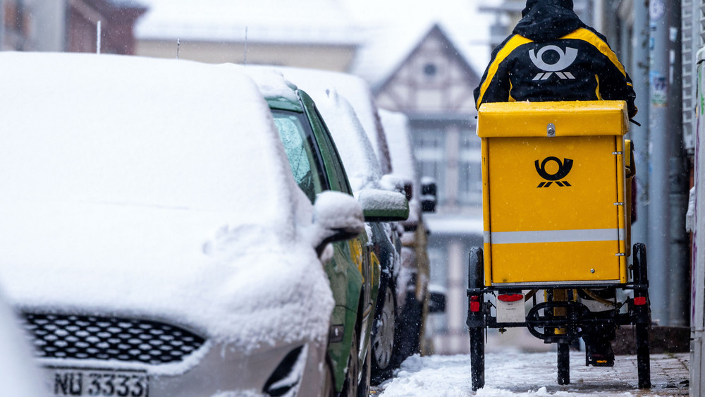 Eine Briefträgerin ist mit ihrem Fahrrad bei starkem Schneefall unterwegs. Künftig soll die Deutsche Bahn mehr Zeit bei der Briefzustellung bekommen.
