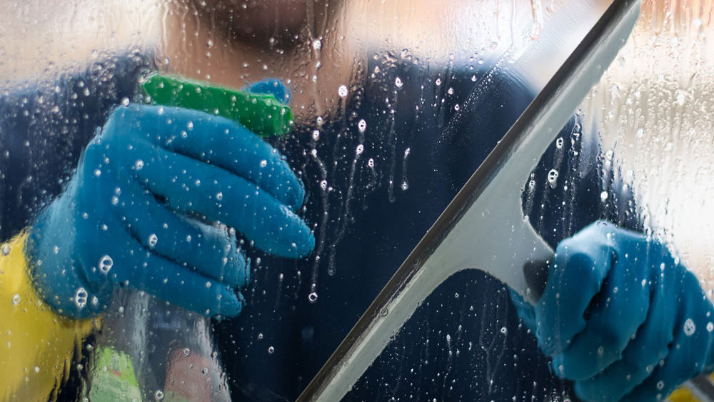 Fenster putzen ohne Streifen: Ein Mann putzt eine Fensterscheibe mit Gummihandschuhen, Reiniger und einem Abzieher
